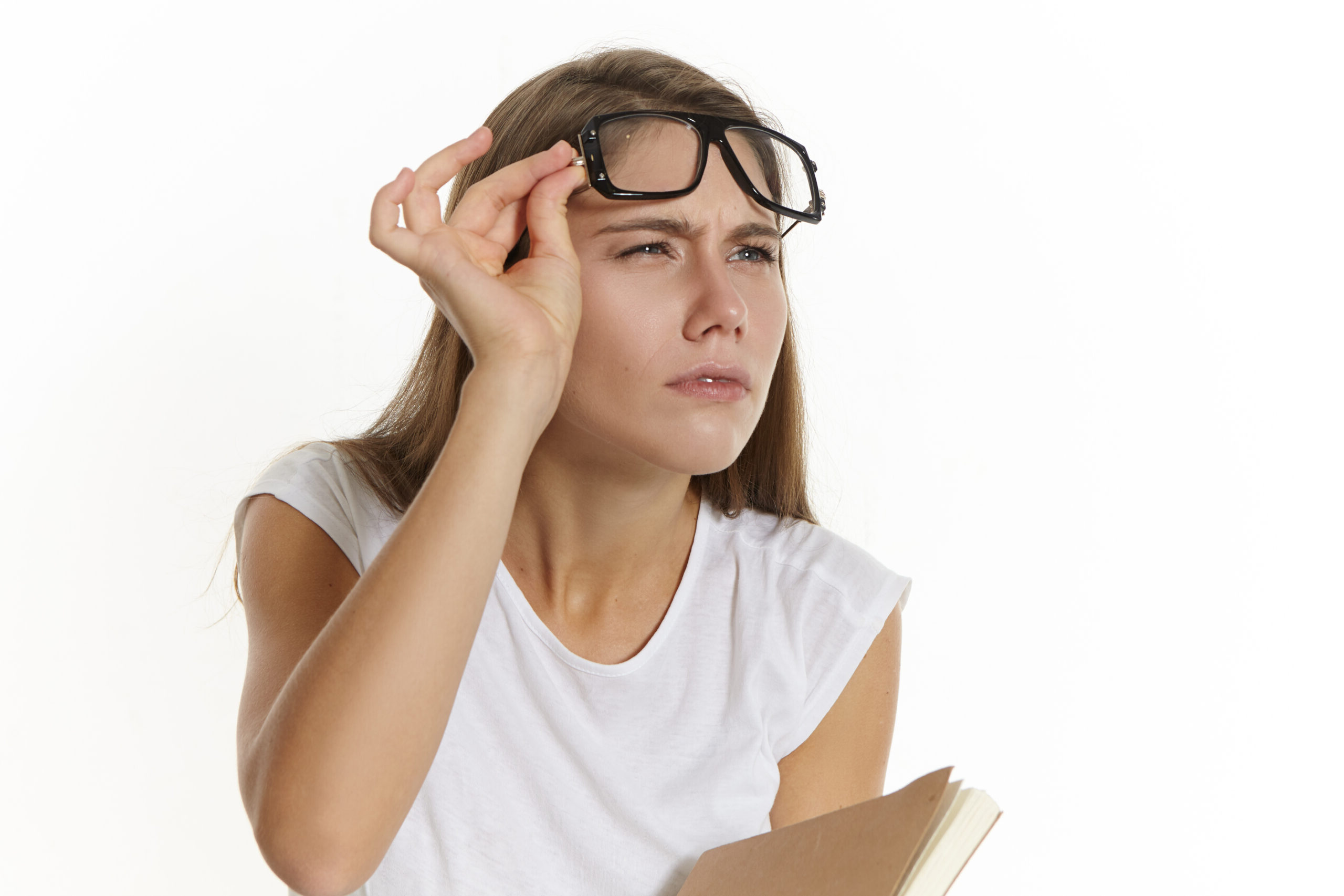 picture of focused serious young caucasian female teacher with textbook, taking off glasses and narrowing eyes, trying to see something clearly. student girl in eyewear posing with diary in studio